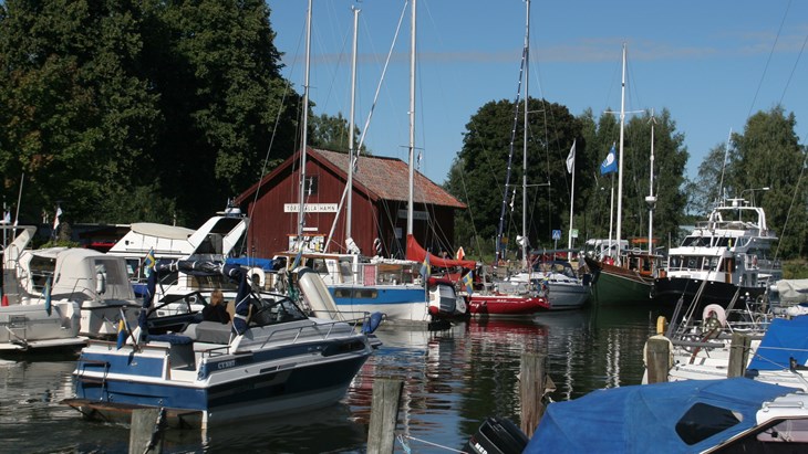 En sommarbild på Torshälla gästhamn. Båtar i vatten och en röd träbyggnad. 