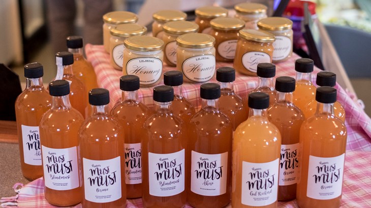 Ett stort antal flaskor med orange/rosa must och burkar med honung på ett bord. 