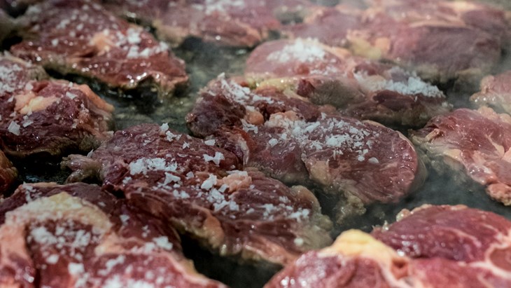 En hand som strör salt över många bitar kött liggandes på ett stekbord.