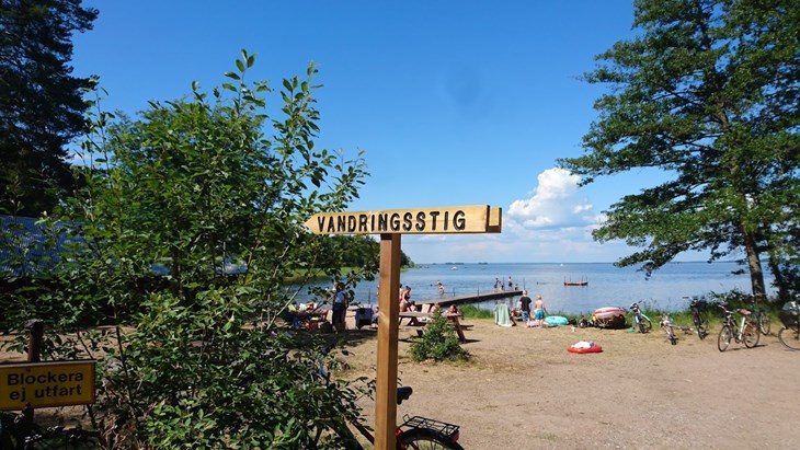 En träskylt med texten Vandringsstig står på en strand intill en sjö.