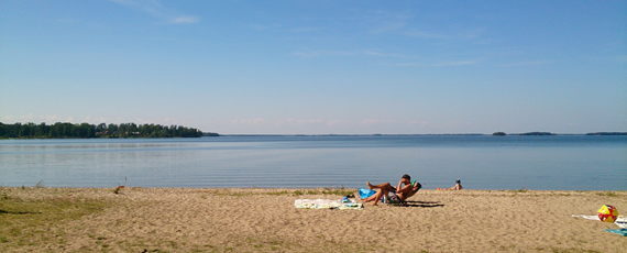 En sandstrand med inslag av gräs intill en sjö. En person sitter i en solstol på stranden.