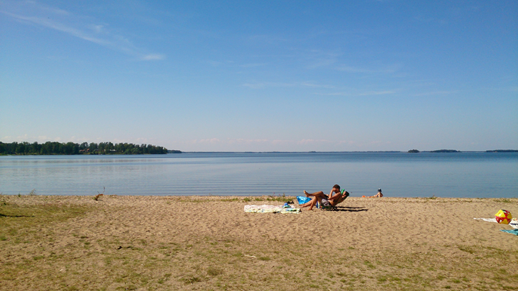 En sandstrand med inslag av gräs intill en sjö. En person sitter i en solstol på stranden.