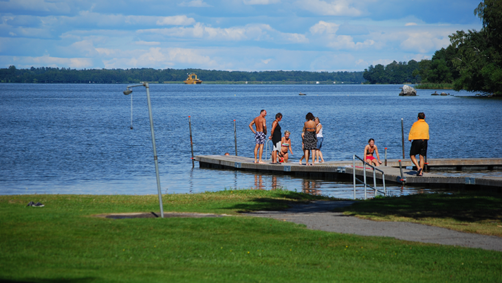 Personer i badkläder står på en brygga vid en sjö.