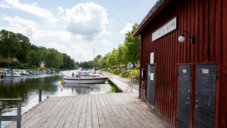 Sommarbild av en brygga med båtar angjorda. Röd träbyggnad med texten Torshälla hamn till höger i bild.