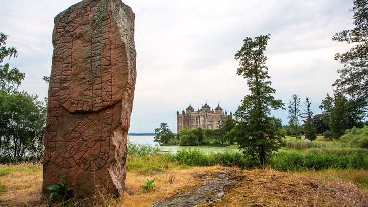 En stor rödbrun sten med runskrift. Ett stort slott som tornar upp sig i bakgrunden.