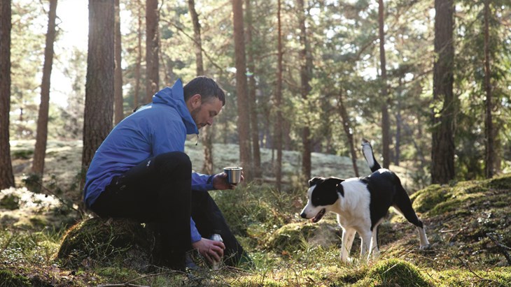 En man i blå jacka som håller en termos med kaffe i en soldränkt skogsdunge. Han har en svartvit minder hund med sig. 
