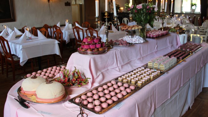 Ett långbord med rosa duk fullt av rosa och vita bakverk. Flera dukade bord med vita dukar runtomkring. 